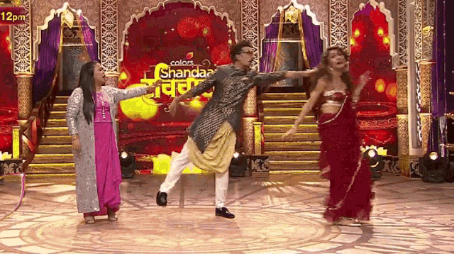 a man and two women are dancing on a stage in front of a banner that says colors shandaar