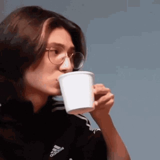 a young man with long hair and glasses is drinking from a cup .