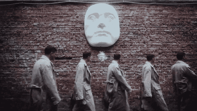 a group of men are walking in front of a brick wall with a large face on it