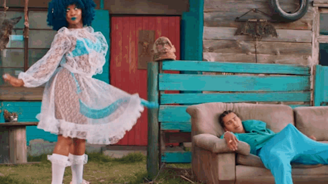 a man in a blue wig is laying on a couch while a woman in a white dress stands behind him