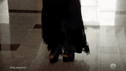 a woman in a black dress and high heels is walking down a tiled floor .