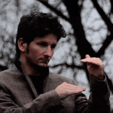 a man in a plaid jacket holds his hands out in front of a tree