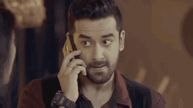 a man with a beard is talking on a cell phone and making a surprised face .