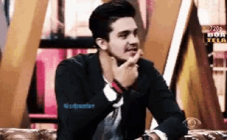 a man in a black jacket is sitting on a couch with his hand to his mouth .