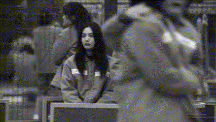 a black and white photo of a woman sitting on a bench with a man walking behind her