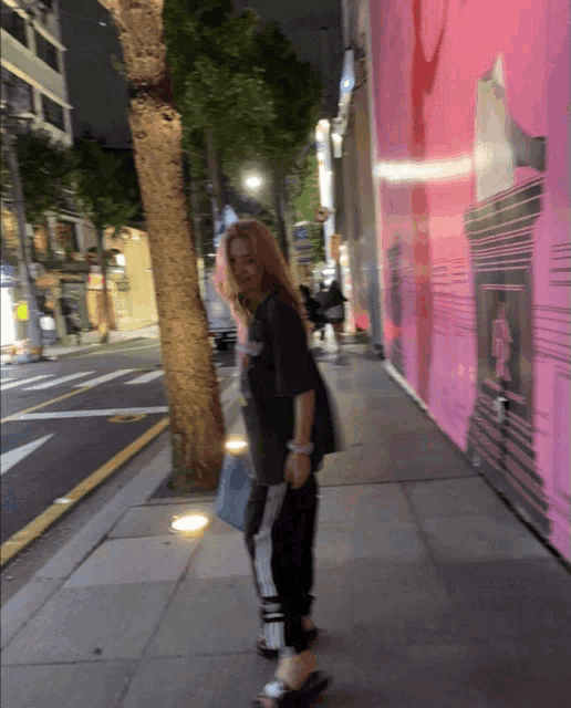 a woman standing on a sidewalk with a pink wall behind her