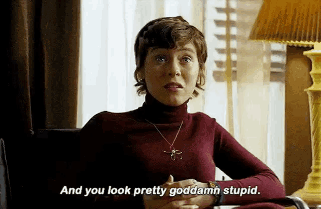 a woman in a red turtleneck sweater is sitting at a table with her hands folded and talking .