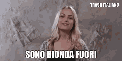 a woman is standing in front of a wall and smiling with the words `` sono bionda fuori '' written above her .