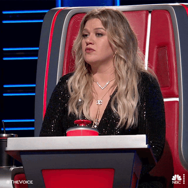 a woman is sitting at a table with a cup of coffee in front of her and the word the voice is on the bottom right