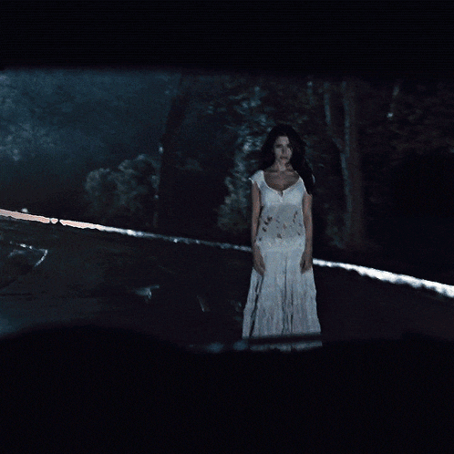 a woman in a white dress stands in the dark in front of a car