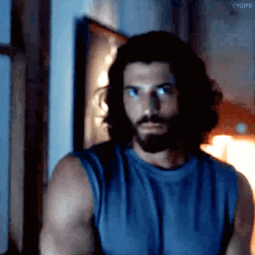 a man with long hair and a beard wearing a blue tank top is standing in a dark room .