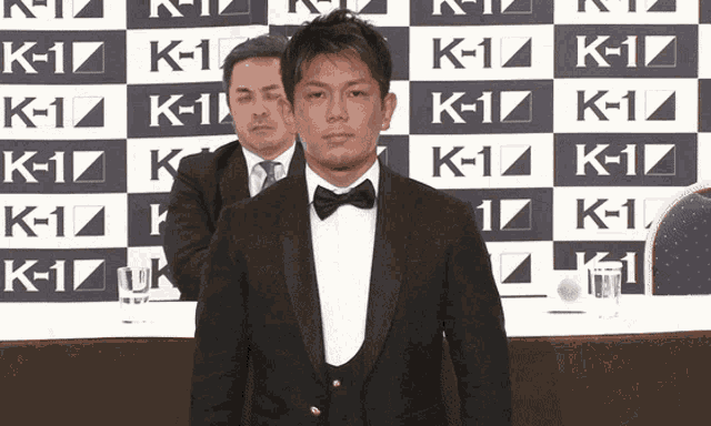 a man in a tuxedo stands in front of a wall that says k- 1