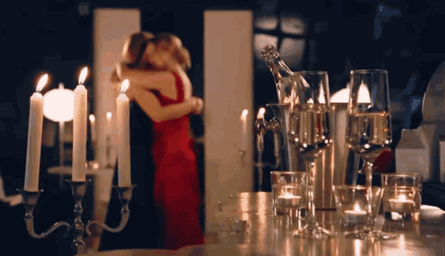 a woman in a red dress is hugging a man in front of a table full of candles and wine glasses