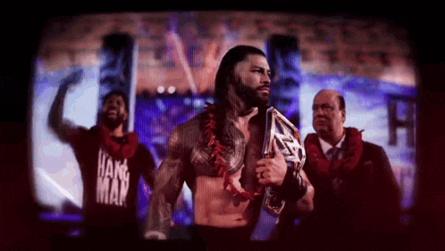 roman reigns is holding a world heavyweight championship belt while standing in front of a group of men .