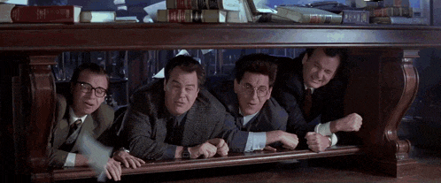 a group of men peeking out from under a table with books on it