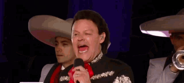 a man in a mariachi outfit is singing into a microphone while two men play trumpets in the background
