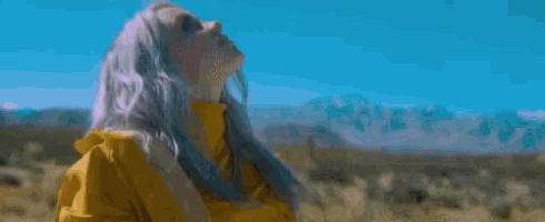 billie eilish is wearing a yellow jacket and standing in the middle of a field .