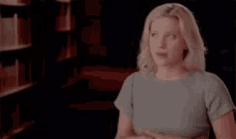 a woman in a blue shirt is standing in front of a bookshelf and talking to someone .