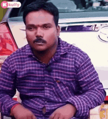a man wearing a purple plaid shirt is sitting in front of a white car .