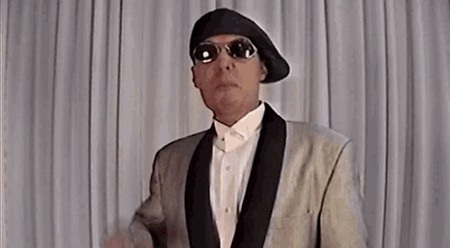 a man wearing a tuxedo and sunglasses stands in front of a white curtain .