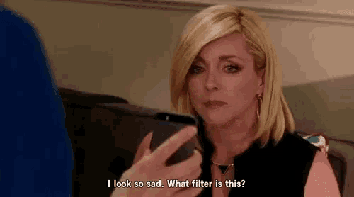 a woman is sitting on a couch looking at her cell phone and saying `` i look so sad what filter is this ? ''