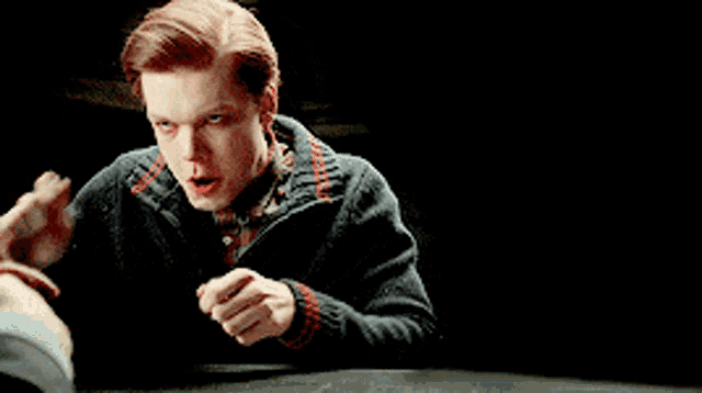 a young man with red hair is sitting at a table with his hands on his chin .