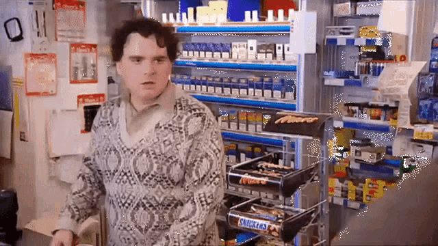 a man in a sweater is standing in front of a shelf with snickers on it .