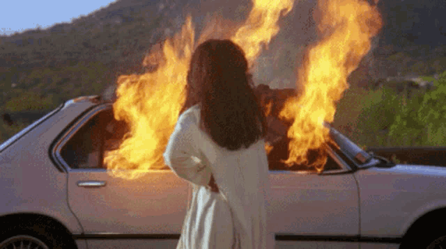 a woman is standing in front of a white car that is on fire .
