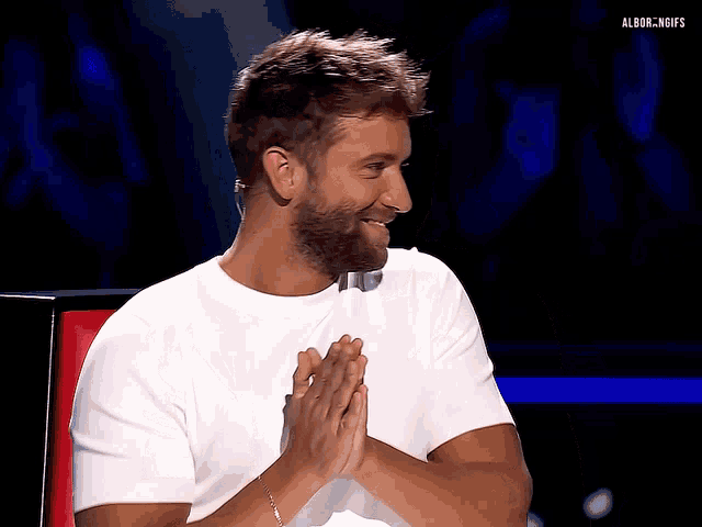 a man with a beard wearing a white t-shirt is sitting with his hands folded in prayer