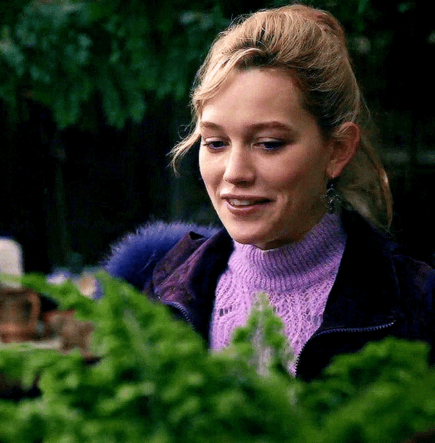 a woman wearing a purple sweater and a purple jacket is looking at a plant .