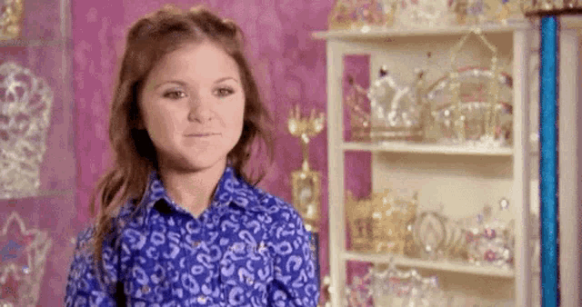 a young girl in a blue shirt is standing in front of a shelf filled with crowns and tiaras .