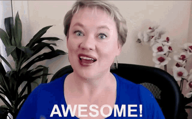 a woman in a blue shirt says awesome in front of a plant