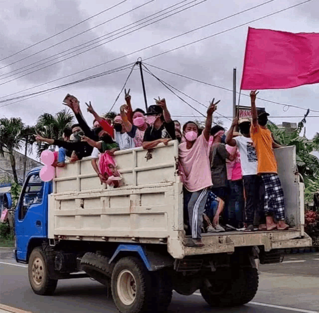 a group of people are riding on the back of a truck with a pink flag in their hand
