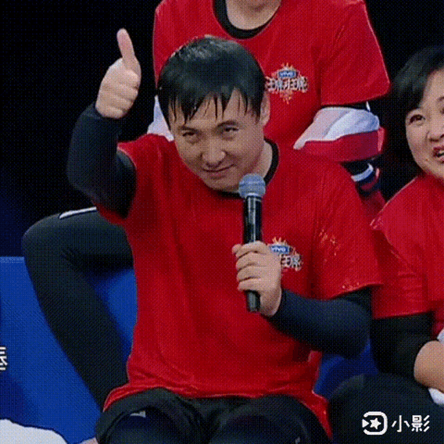 a man in a red shirt is holding a microphone and giving a thumbs up sign .