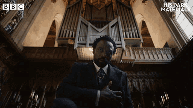 a man in a suit and tie is sitting in front of an organ with the words his dark materials on the bottom right