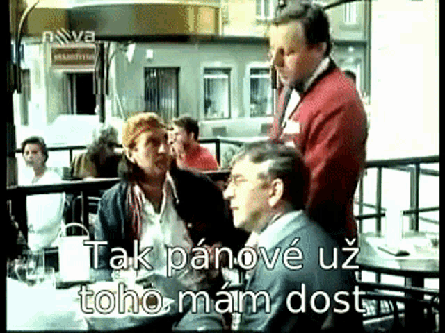 a group of people are sitting at a table with a man in a red jacket standing behind them ..