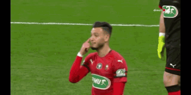 a soccer player wearing a red puma jersey is pointing at his nose