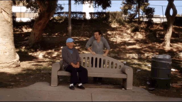 two men sit on a bench in a park talking
