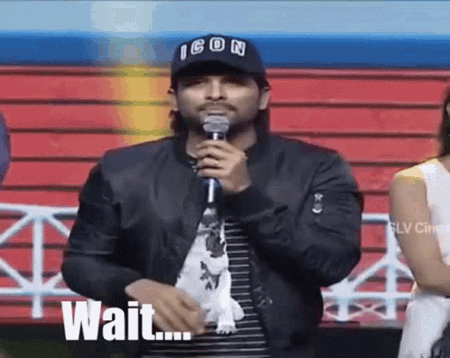 a man in an icon hat is holding a microphone and says wait