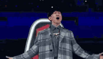 a man in a plaid jacket is standing on a stage with his arms outstretched and screaming .