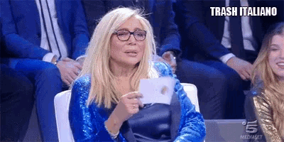 a woman wearing glasses and a blue sequined dress is sitting in front of a crowd .