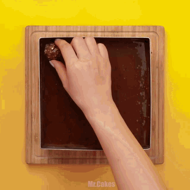 a person 's hand is holding a piece of chocolate on top of a wooden cutting board that says mr.cakes