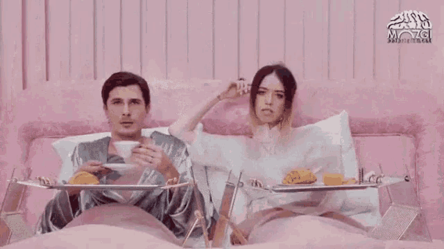 a man and a woman are sitting in a bed with trays of food .