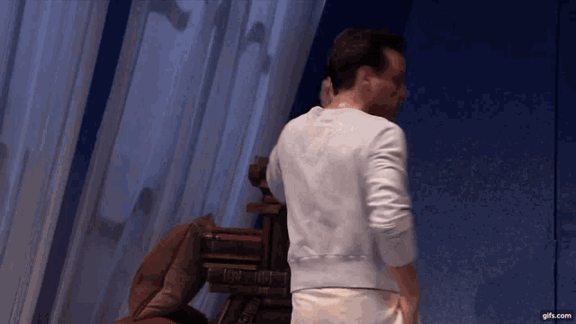 a man in a white sweater is standing in front of a bookshelf with books on it .