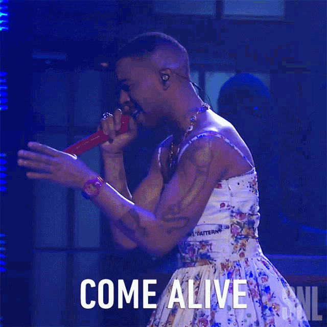 a man in a floral dress singing into a microphone with the words come alive written below him