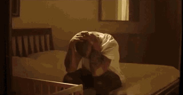 a man is sitting on a bed with his head in his hands and a baby in the background .