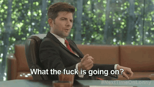 a man in a suit and tie is sitting at a desk and says what the fuck is going on