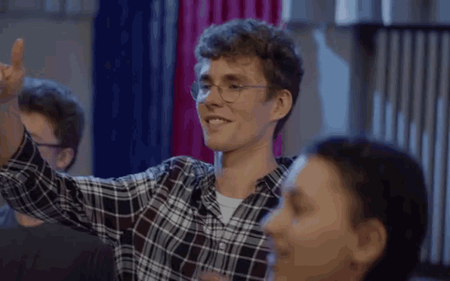 a man wearing glasses and a plaid shirt is smiling and raising his hand in the air