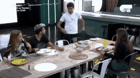 a group of people are sitting at a table with a man standing in the background