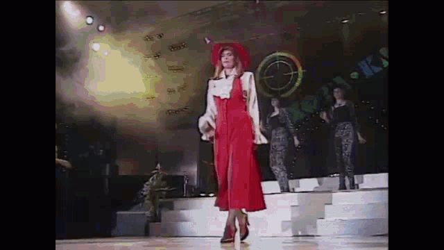 a woman in a red dress and red hat is walking down a stage .